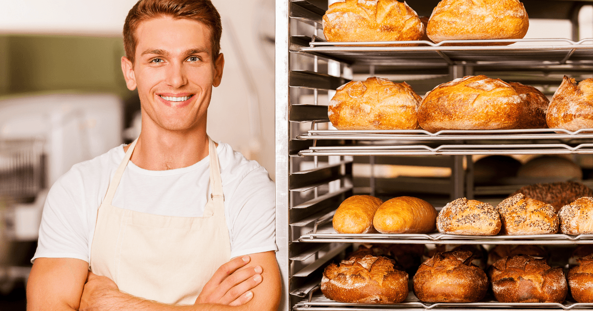 Handsome young baker in apron keeping arms crossed and smiling while leaning at the tray with fresh baked bread